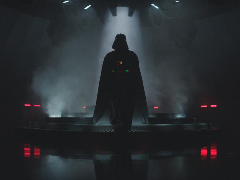 Fimausschnitt zeigt Darth Vader in einer großen, dunklen Halle; er läuft auf die Kamera zu und geht eine Treppe mit wenigen Stufen hinunter.