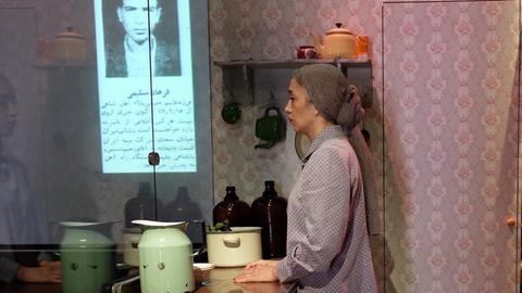 Eine Frau schaut ins Leere. Sie steht an einer Küchenarbeitsplatte, im Hintergrund eine geblümte Tapete. Sie trägt ein Kopftuch und wirkt gedankenverloren bis traurig. Im Hintergrund das Bild eines Mannes, darunter arabische Schriftzeichen. 
