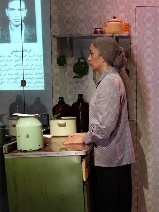 Eine Frau schaut ins Leere. Sie steht an einer Küchenarbeitsplatte, im Hintergrund eine geblümte Tapete. Sie trägt ein Kopftuch und wirkt gedankenverloren bis traurig. Im Hintergrund das Bild eines Mannes, darunter arabische Schriftzeichen. 