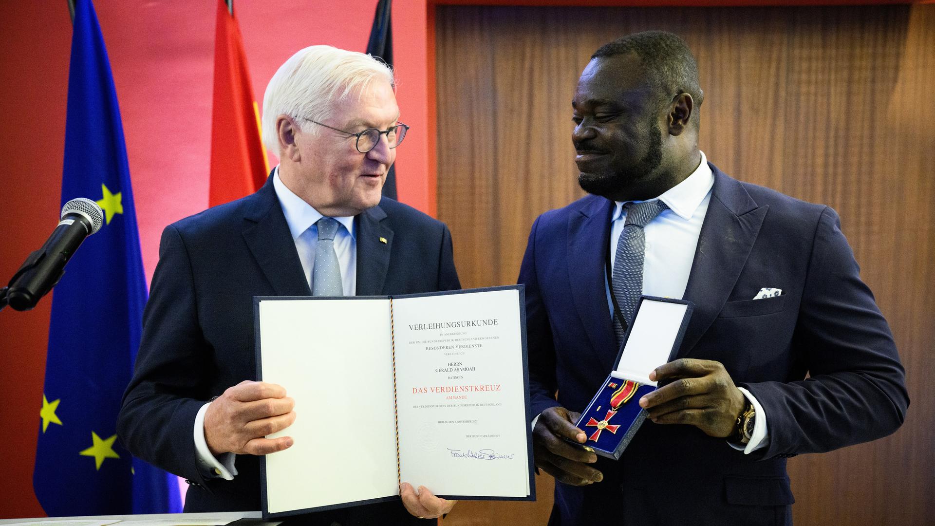 Bundespräsident Frank-Walter Steinmeier steht neben Gerald Asamoah. Asamoa hält das Bundesverdienstkreuz in der Hand. Steinmeier trägt die dazugehörige Urkunde.  Bundespräsident Frank-Walter Steinmeier steht neben Gerald Asamoah. Asamoa hält das Bundesverdienstkreuz in der Hand. Steinmeier trägt die dazugehörige Urkunde.
