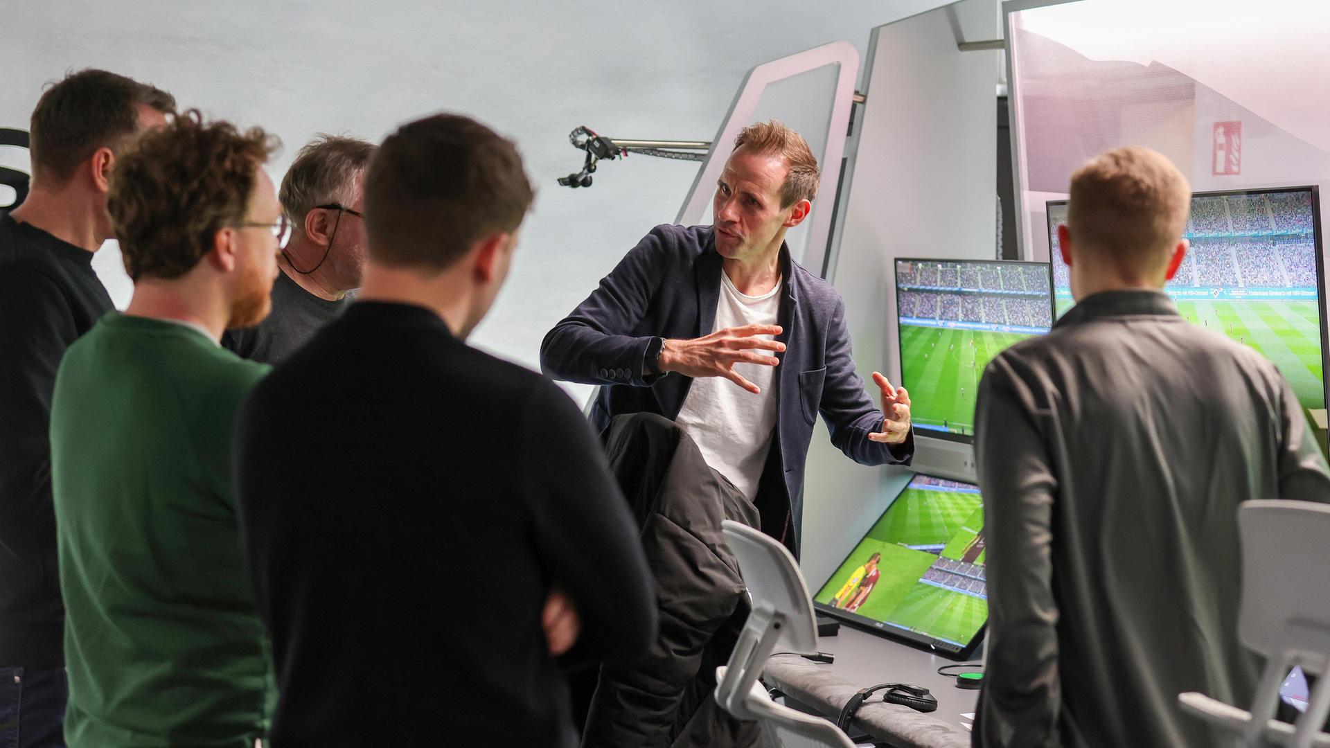Eine Personengruppe steht dem Betrachter mit dem Rücken zugewandt vor einer Wand mit mehreren Monitoren, auf denen ein Fußballfeld im Stadion zu sehen ist. Eine Person erklärt gestikulierend das Prozedere. Eine Personengruppe steht dem Betrachter mit dem Rücken zugewandt vor einer Wand mit mehreren Monitoren, auf denen ein Fußballfeld im Stadion zu sehen ist. Eine Person erklärt gestikulierend das Prozedere.