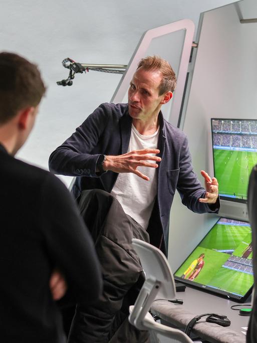 Eine Personengruppe steht dem Betrachter mit dem Rücken zugewandt vor einer Wand mit mehreren Monitoren, auf denen ein Fußballfeld im Stadion zu sehen ist. Eine Person erklärt gestikulierend das Prozedere.