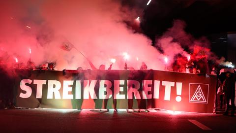 Beschäftigte tragen in der Dunkelheit ein Plakat mit der Aufschrift "Streikbereit!". Im Hintergrund Rauch und rötliches Licht.