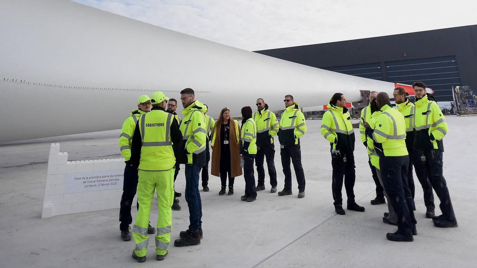 Ein Teil eines Windrades liegt vor einer schwarzen Halle bei Siemens Gamesa in Le Havre in Frankreich. Davor stehen ein Dutzend Arbeiter in gelben Arbeitsjacken. 