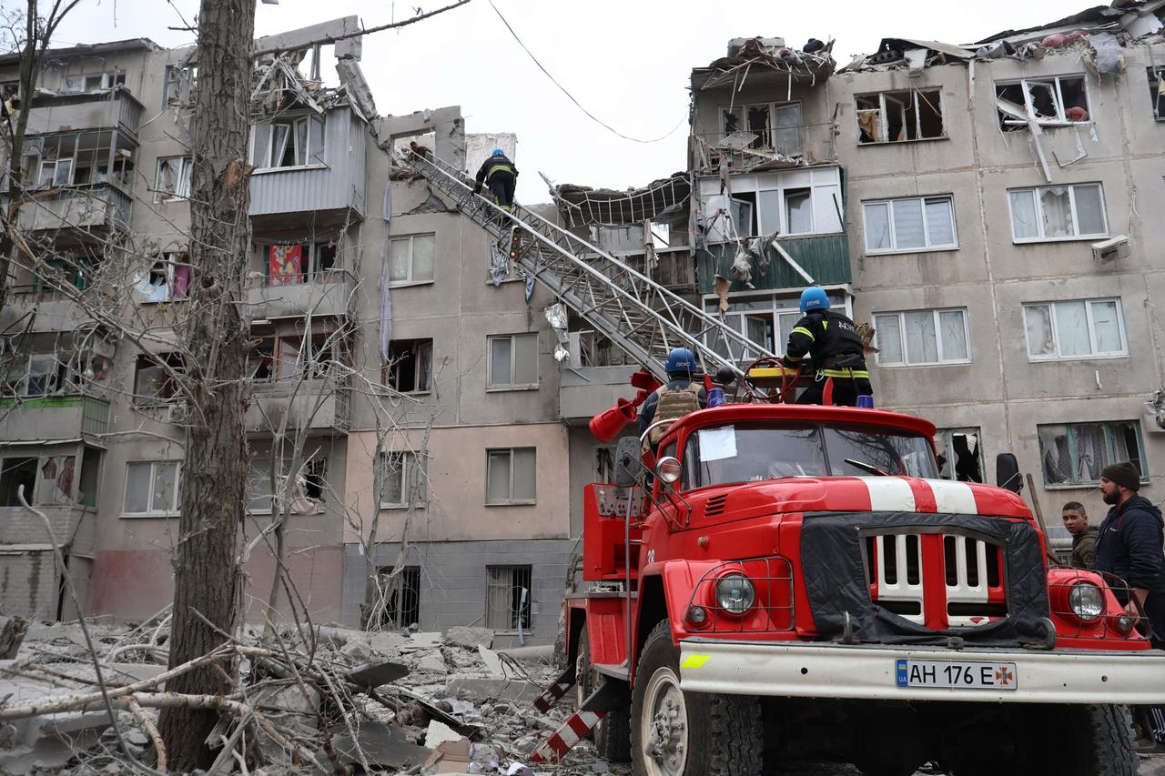 An einem zerbombten Mehrfamilienhaus steht ein Feuerwehrwagen. Feuerwehrleute auf Drehleitern suchen nach Überlebenden.