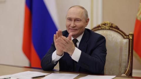 Putin sitzt an einem Tisch und klatscht in die Hände. Hinter ihm steht eine russische Nationalfahne.