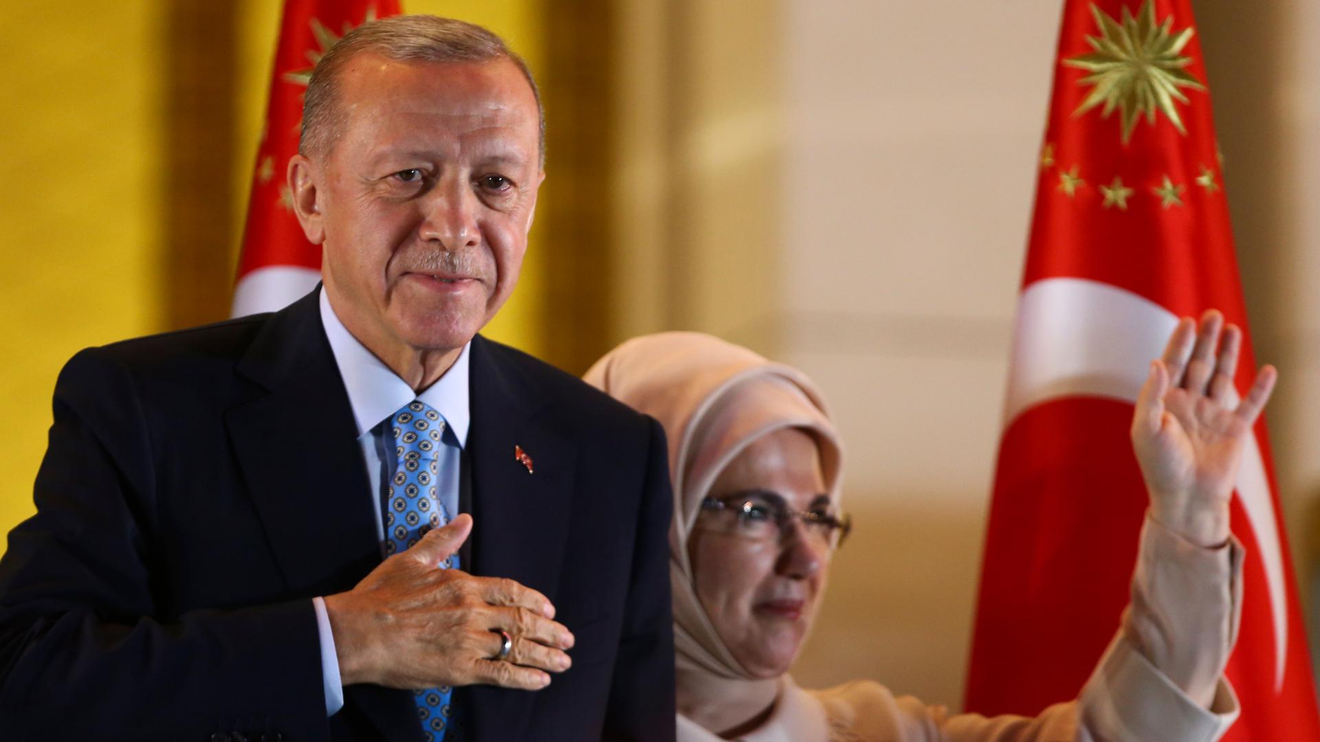Das Foto zeigt den türkischen Präsidenten Erdogan. Er hält sich eine Hand vor die Brust. Daneben steht seine Frau. Sie winkt.