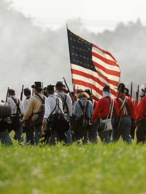 Ein Reenactment des Amerikanischen Bürgerkriegs auf einer Wiese. Eine Gruppe von Männern läuft dicht gedrängt, in der Mitte der Gruppe trägt einer die US-Flagge.