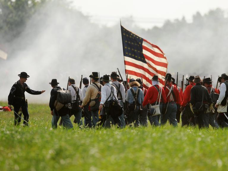 Ein Reenactment des Amerikanischen Bürgerkriegs auf einer Wiese. Eine Gruppe von Männern läuft dicht gedrängt, in der Mitte der Gruppe trägt einer die US-Flagge.