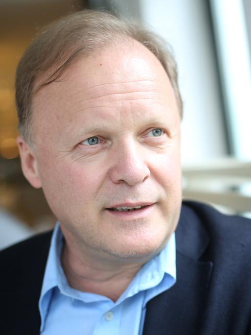 Stephan Werhahn sitzt auf einer Bank in einem Café und schaut aus dem Fenster. Er trägt ein hellblaues Hemd zum dunkelblauen Anzug.