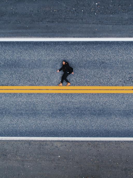 Luftaufnahme einer menschlichen Gestalt auf dem Asphalt liegend. Es wirkt auf den ersten Blick als würde die Person aufrecht laufen, statt flach auf dem Boden zu liegen. Unter den Füßen eine gelbe, horizontale Linie.