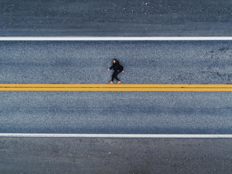 Luftaufnahme einer menschlichen Gestalt auf dem Asphalt liegend. Es wirkt auf den ersten Blick als würde die Person aufrecht laufen, statt flach auf dem Boden zu liegen. Unter den Füßen eine gelbe, horizontale Linie.
