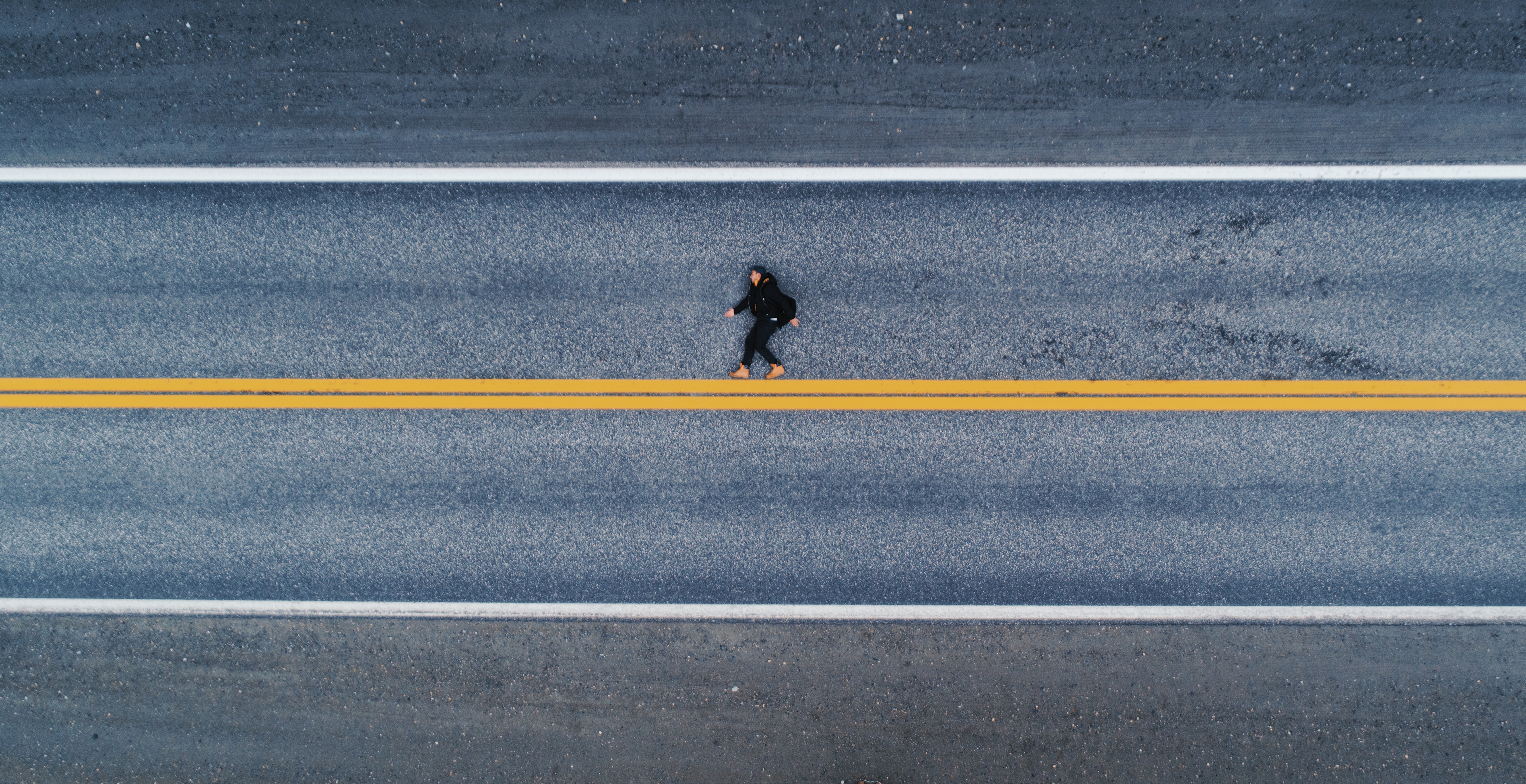 Luftaufnahme einer menschlichen Gestalt auf dem Asphalt liegend. Es wirkt auf den ersten Blick als würde die Person aufrecht laufen, statt flach auf dem Boden zu liegen. Unter den Füßen eine gelbe, horizontale Linie.