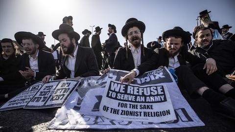 Mehrere Männer mit den markanten schwarzen Hüten und den Schläfenloclemn stehen nebeneinander und halten Plakate mit Slogans gegen ihre Einberufung in der Hand. 