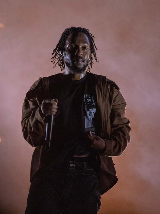 Der Rapper Kendrick Lamar steht auf einer Bühne, im Hintergrund sind Flammen zu sehen.
