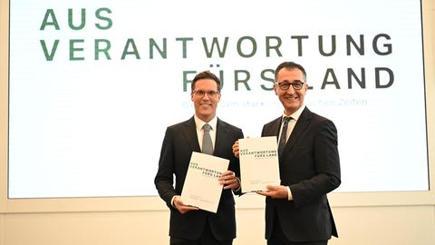 Cem Özdemir und Manuel Hagel halten jeweils den Koalitionsvertrag in den Händen und lächeln in die Kamera. 