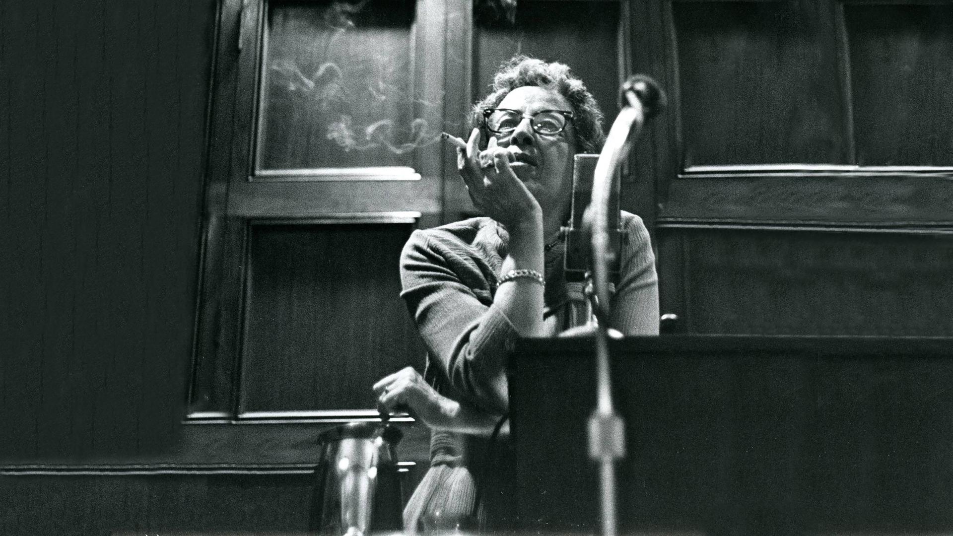 Filmstill aus "Hannah Arendt - Die Pflicht zum Ungehorsam" in dem Hannah Arendt Zigarette rauchend sitzend an einem Tisch lehnt. 