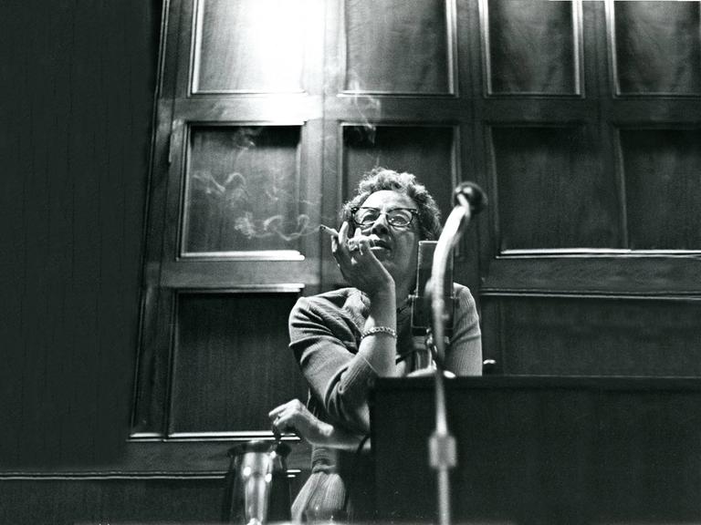 Filmstill aus "Hannah Arendt - Die Pflicht zum Ungehorsam" in dem Hannah Arendt Zigarette rauchend sitzend an einem Tisch lehnt. 
