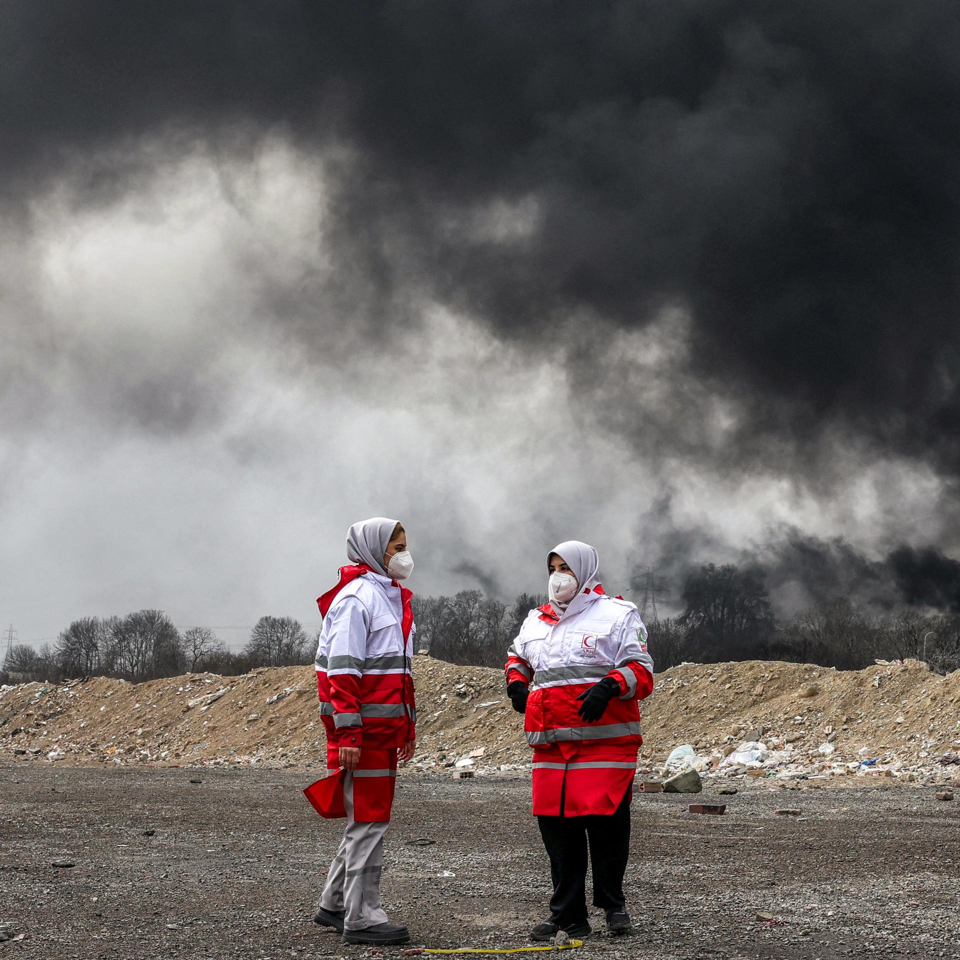 Frauen des iranischen Roten Halbmonds, erkennbar durch die rot-weiße Kleidung, stehen vor dichten schwarzen Rauchwolken eines Brandes nach einem nächtlichen Luftangriff auf eine Ölraffinerie im Nordwesten Teherans. Sie tragen Mundschutz.
