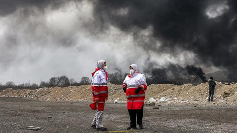 Frauen des iranischen Roten Halbmonds, erkennbar durch die rot-weiße Kleidung, stehen vor dichten schwarzen Rauchwolken eines Brandes nach einem nächtlichen Luftangriff auf eine Ölraffinerie im Nordwesten Teherans. Sie tragen Mundschutz.