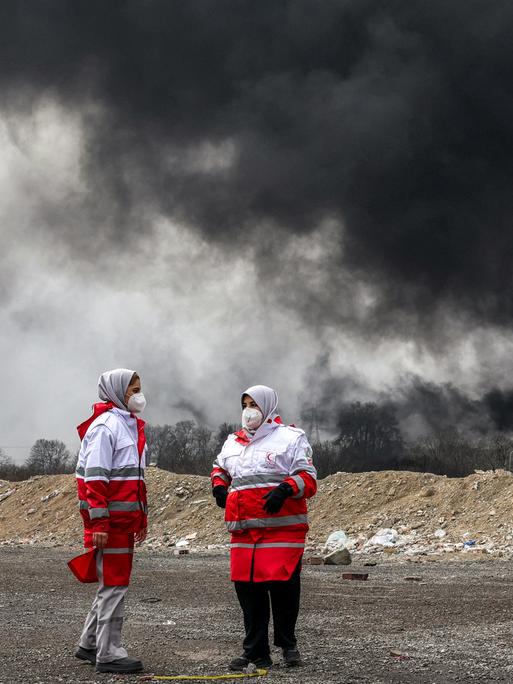 Frauen des iranischen Roten Halbmonds, erkennbar durch die rot-weiße Kleidung, stehen vor dichten schwarzen Rauchwolken eines Brandes nach einem nächtlichen Luftangriff auf eine Ölraffinerie im Nordwesten Teherans. Sie tragen Mundschutz.