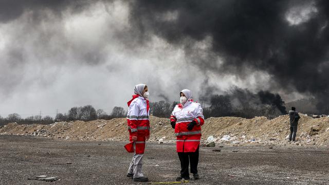 Frauen des iranischen Roten Halbmonds, erkennbar durch die rot-weiße Kleidung, stehen vor dichten schwarzen Rauchwolken eines Brandes nach einem nächtlichen Luftangriff auf eine Ölraffinerie im Nordwesten Teherans. Sie tragen Mundschutz.