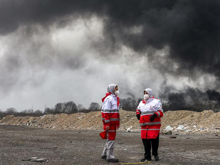 Frauen des iranischen Roten Halbmonds, erkennbar durch die rot-weiße Kleidung, stehen vor dichten schwarzen Rauchwolken eines Brandes nach einem nächtlichen Luftangriff auf eine Ölraffinerie im Nordwesten Teherans. Sie tragen Mundschutz.