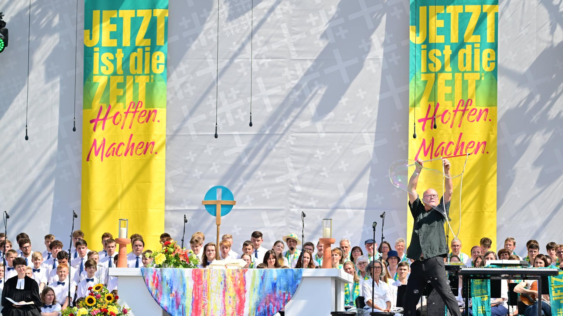 Abschlussgottesdienst des Evangelischen Kirchentags in Nürnberg. Ein Seifenblasenkünstler steht vor einem Altar auf der Bühne. Im Hintergrund sind zwei Banner mit der Aufschrift "jetzt ist die Zeit. Hoffen. Machen"