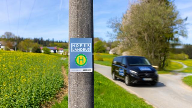 Ein Haltestellenschild mit der Beschriftung "Hofer Landbus", daneben ein Auto in Bewegung