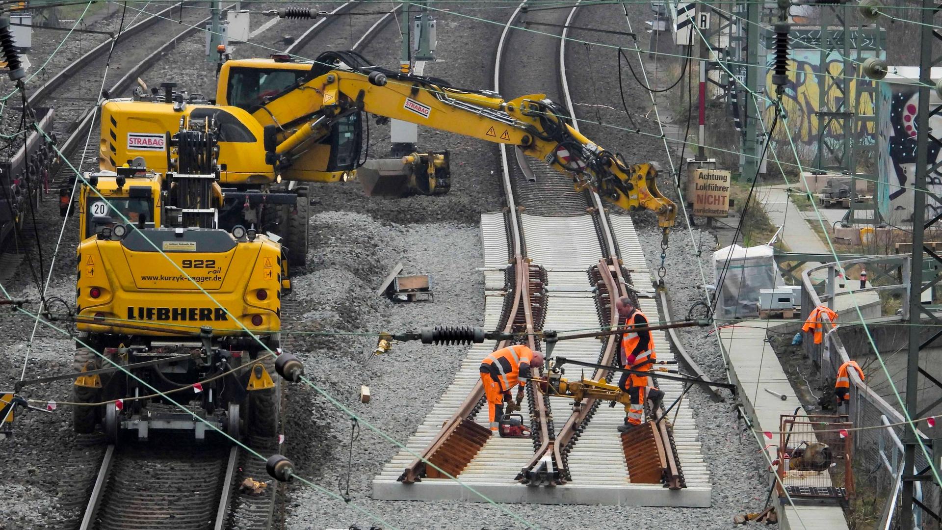 Auf einer Schiene der Deutschen Bahn laufen Sanierungsarbeiten - zu sehen ist ein Bagger und mehrere Bauarbeiter, die an den Gleisen arbeiten.