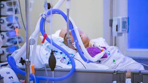 Ein Mensch liegt in lilafarbener Kleidung in einem Bett. Sein Gesicht ist von Schläuchen und technischem Gerät verdeckt. 