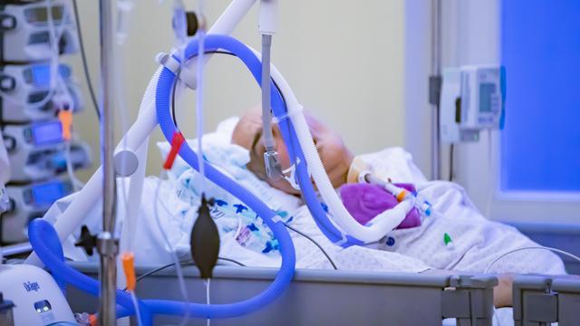 Ein Mensch liegt in lilafarbener Kleidung in einem Bett. Sein Gesicht ist von Schläuchen und technischem Gerät verdeckt. 
