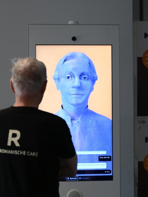 Ein Besucher steht im Käthe-Kollwitz-Museum mit einem mit 3D-Animation und künstlicher Intelligenz hergestellten digitalen Avatar in der Gestalt von Käthe Kollwitz.