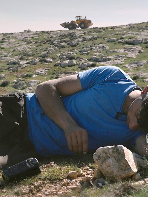 Eine Szene aus dem Film "No Other Land", ein junger Mann liegt auf felsigem Untergrund, dahinter sieht man einen Bagger.
