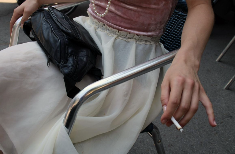 Der sitzende Ausschnitt einer Person im Rock und rosa Oberteil, mit Perlenkette um den Hals und Zigarette in der Hand, eine Tasche auf dem Schoß