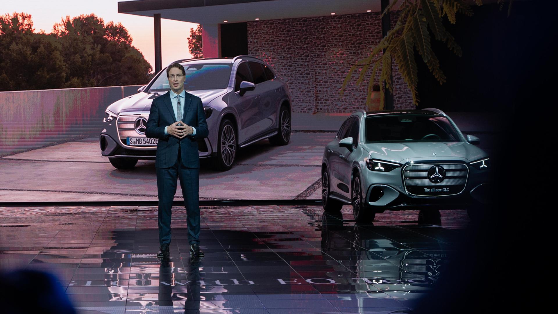 Ola Källenius spricht auf einer Pressekonferenz. Hinter ihm stehen zwei Autos. 