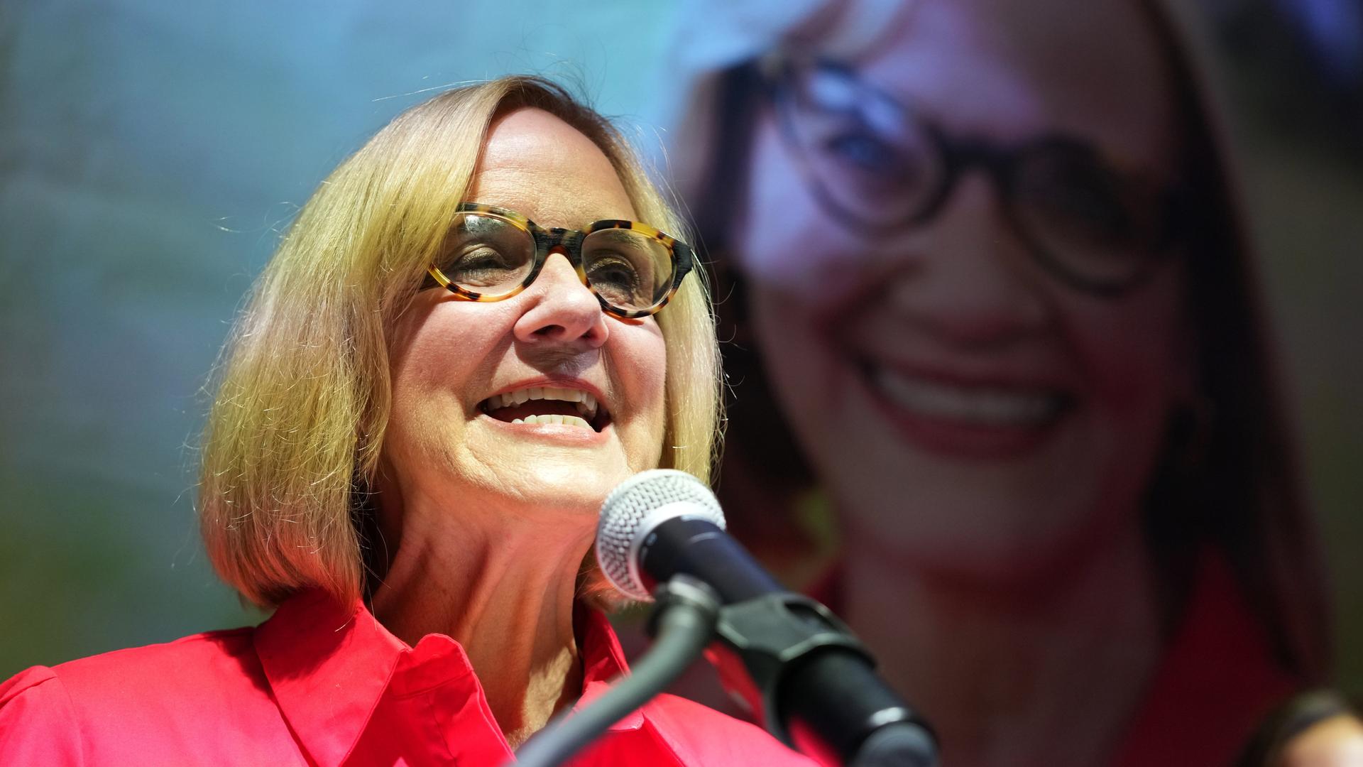 Higgins im roten Hemd und mit Brille spricht lächelnd in ein Mikrofon. Im Hintergrund sieht man sie groß auf einer Videoleinwand.  