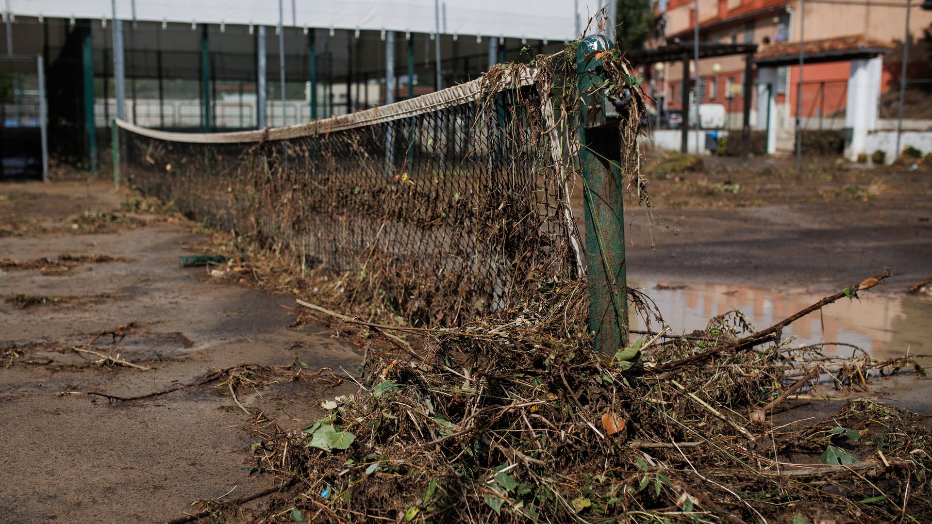 Blick auf einen verwüsteten Tennisplatz in Spanien. Am Netz hängen Sträucher. Der Boden ist mit Schlamm übersät. 