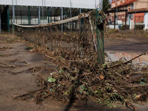 Blick auf einen verwüsteten Tennisplatz in Spanien. Am Netz hängen Sträucher. Der Boden ist mit Schlamm übersät. 