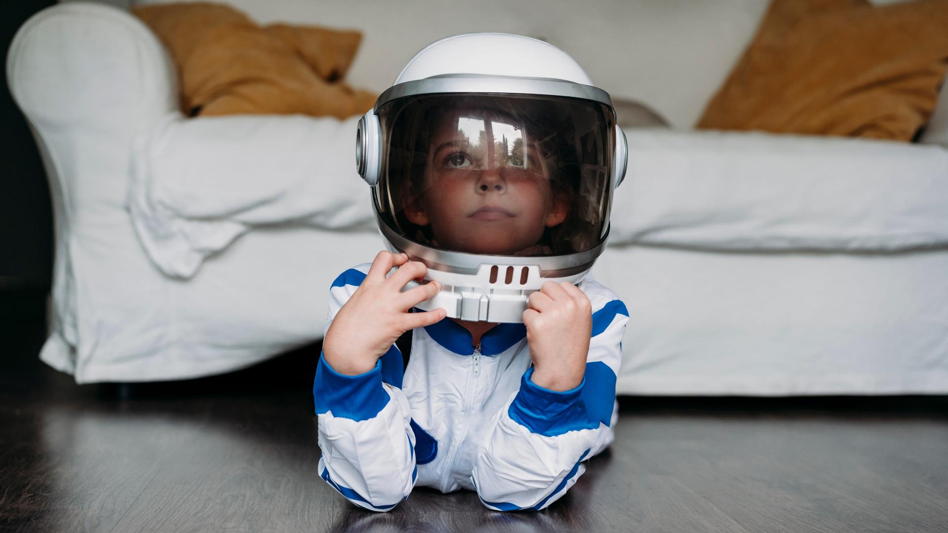 Ein Kind liegt in einem Astronautenkostüm auf dem Boden vor einem Sofa und guckt verträumt.