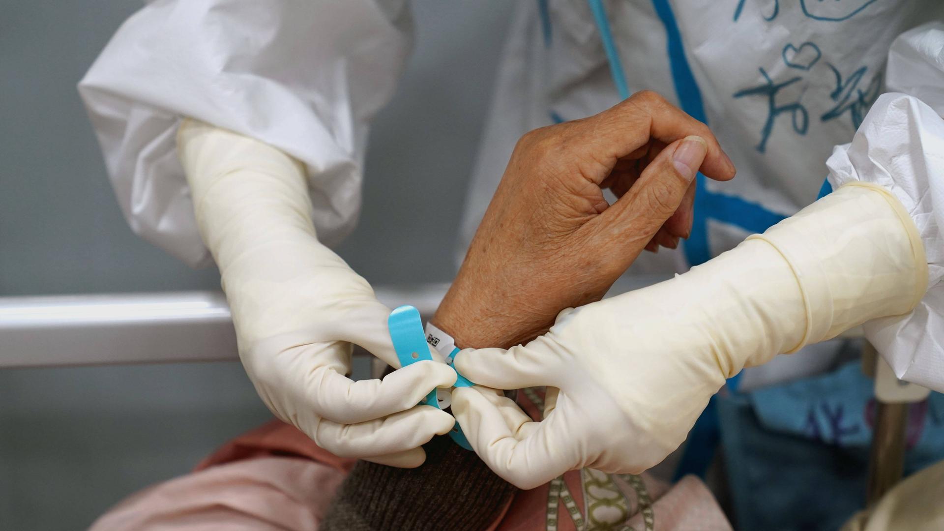 Ein Mensch in weißem Schutzanzug legt an das Handgelenk einer Patientin ein Armband an.