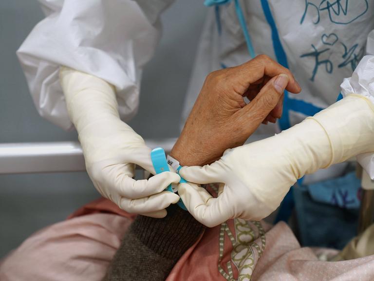 Ein Mensch in weißem Schutzanzug legt an das Handgelenk einer Patientin ein Armband an.
