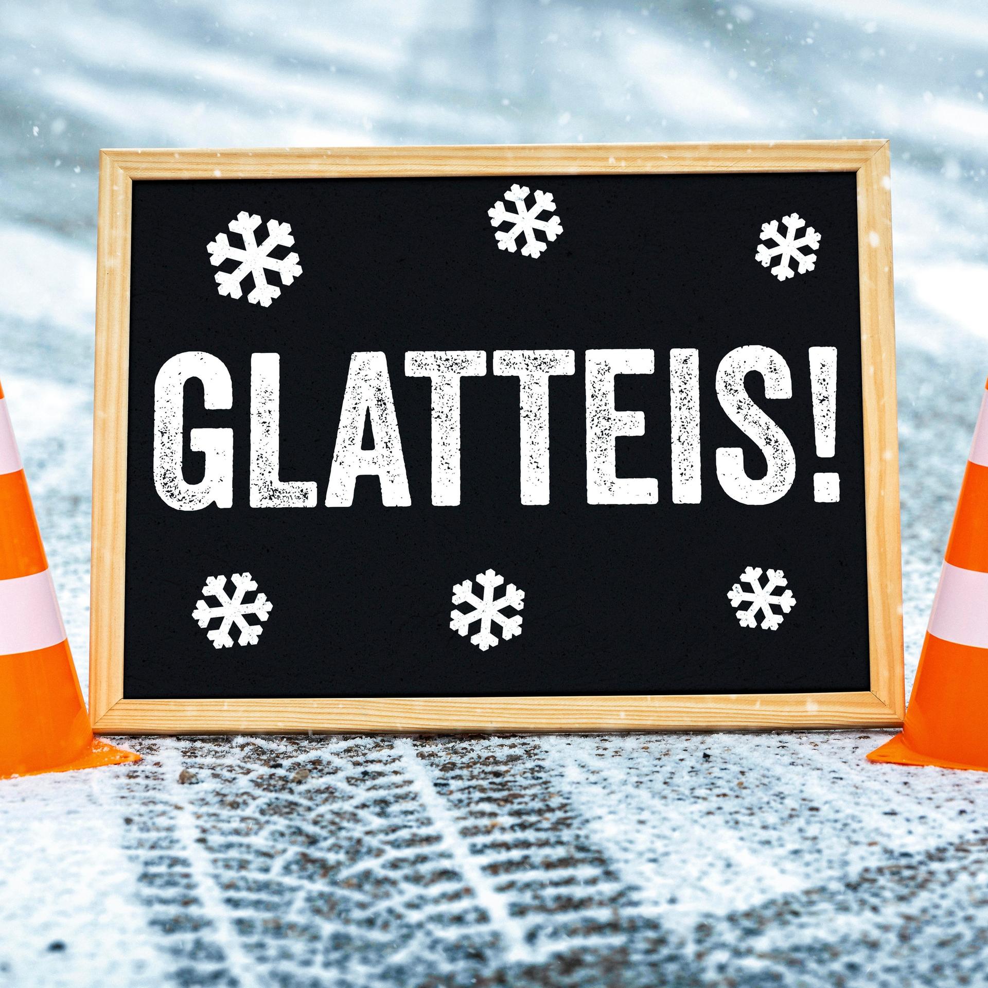 Symbolbild Glatteis: Zwischen zwei orange-weißen Pylonen auf einer verschneiten Straße ist ein Schild, auf dem "Glatteis" steht.