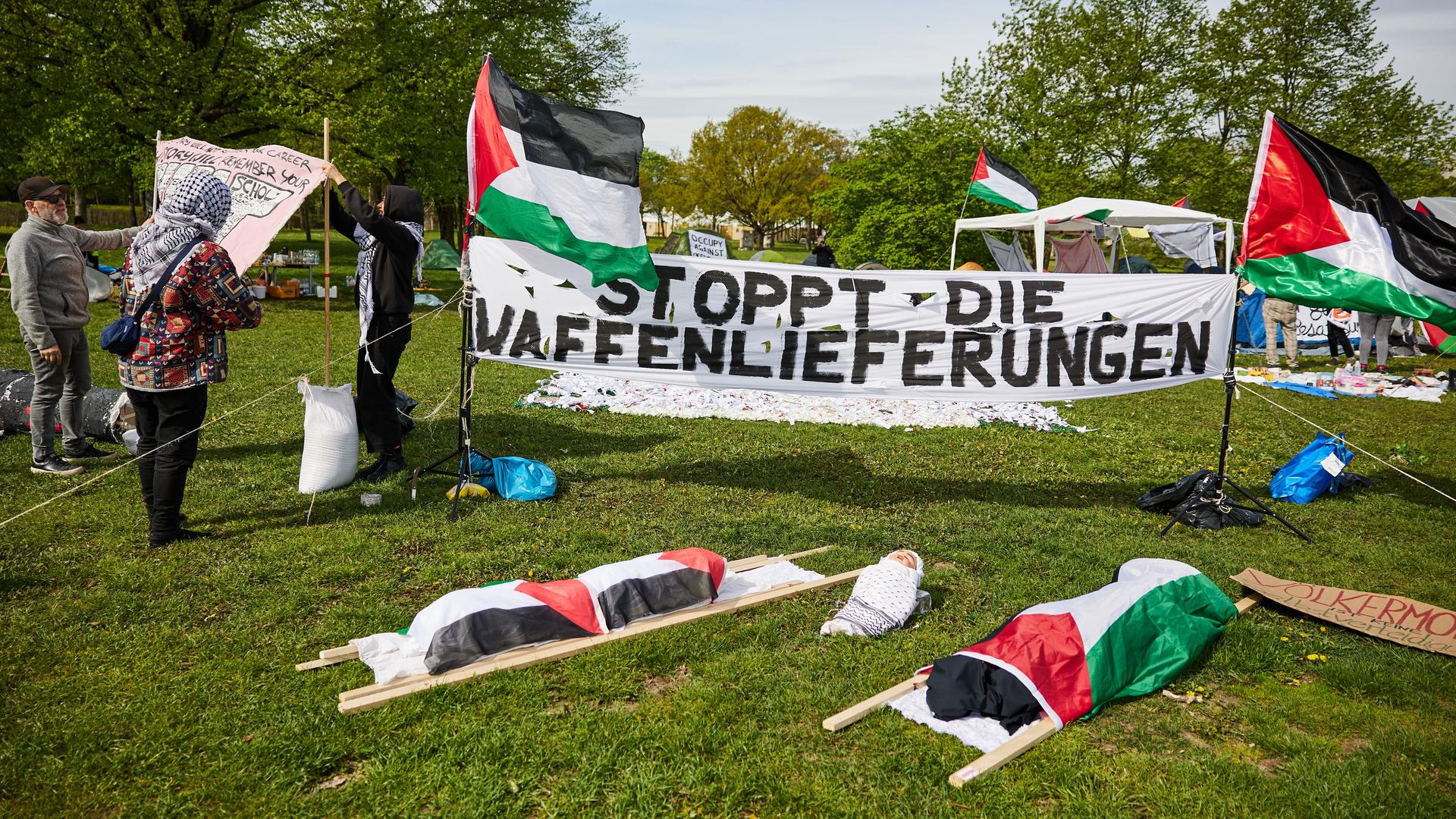 Auf der Wiese vor dem Reichstagsgebäude sieht man ein Banner mit der Aufschrift "Stoppt die Waffenlieferungen". Daneben sind zwei palästinenische Fahnen und zwei Toten-Bahren, die mit palästinensischen Fahnen umwickelt sind.