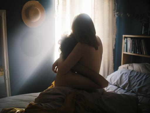 Ein Paar umarmt sich in einem warm beleuchteten Schlafzimmer, in dem Sonnenlicht durch die Vorhänge fällt. 