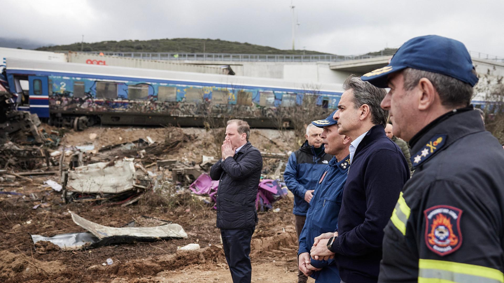 Griechenlands Verkehrsminister Karamanlis und Ministerpräsident Mitsotakis in einer Gruppe von Männern am Unglücksort. Karamanlis schlägt die Hände vors Gesicht. Griechenlands Verkehrsminister Karamanlis und Ministerpräsident Mitsotakis in einer Gruppe von Männern am Unglücksort. Karamanlis schlägt die Hände vors Gesicht.