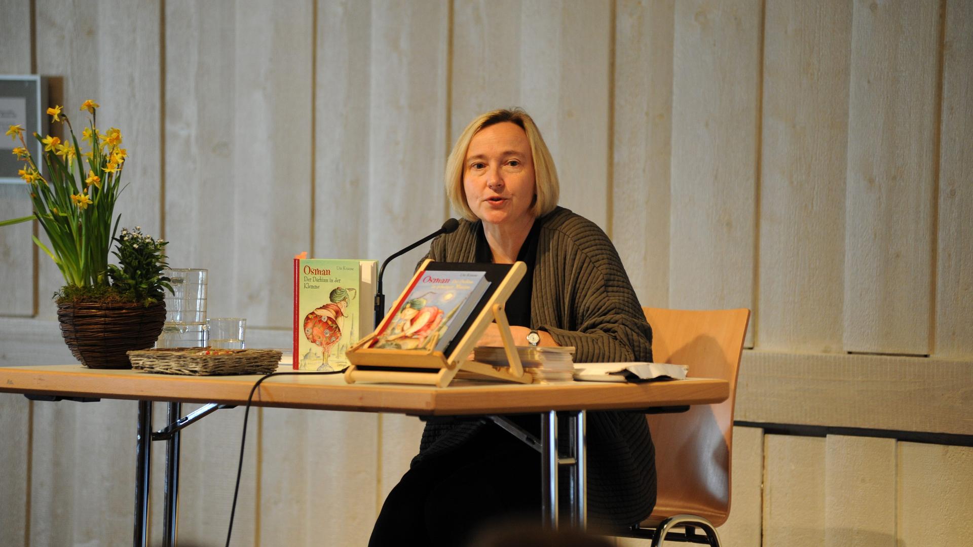 Ute Krause, deutsche Schriftstellerin und Illustratorin von Kinder- und Jugendbüchern bei einer Lesung. Sie sitzt an einem Tisch, trägt eine dunkle Jacke und vor ihr liegen und stehen einige ihrer Bücher.