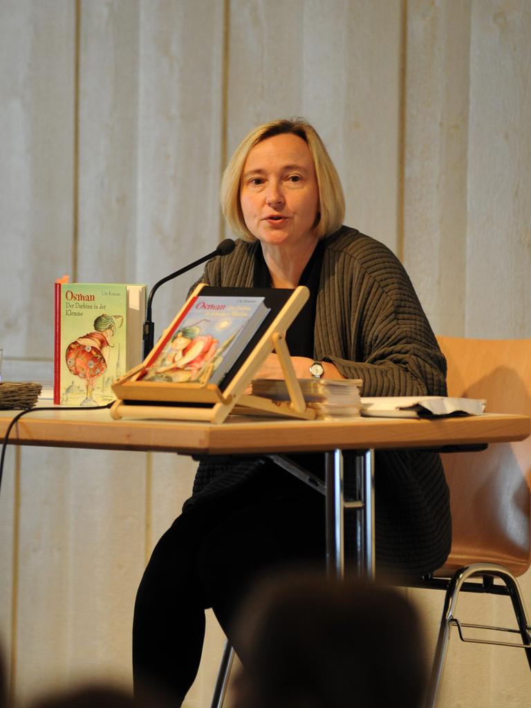 Ute Krause, deutsche Schriftstellerin und Illustratorin von Kinder- und Jugendbüchern bei einer Lesung. Sie sitzt an einem Tisch, trägt eine dunkle Jacke und vor ihr liegen und stehen einige ihrer Bücher.