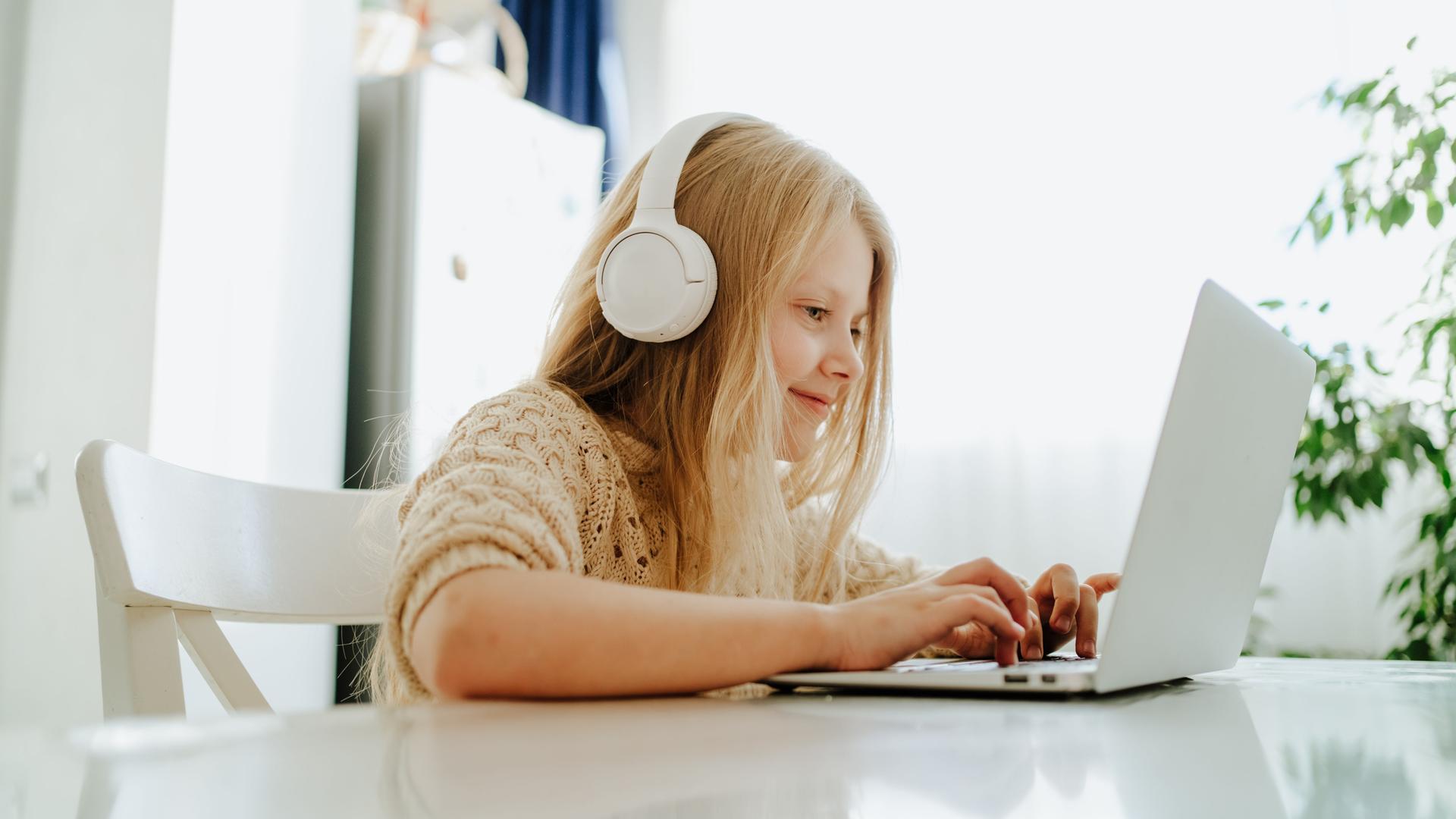 Ein Mädchen mit weißen Kopfhörern sitzt am Tisch vor einem Laptop und schreibt. Ein Mädchen mit weißen Kopfhörern sitzt am Tisch vor einem Laptop und schreibt.