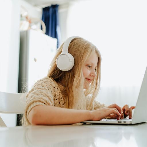 Ein Mädchen mit weißen Kopfhörern sitzt am Tisch vor einem Laptop und schreibt.